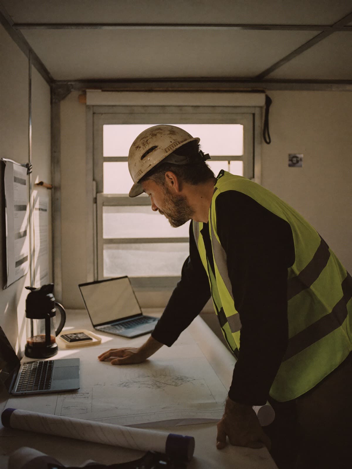 Construction superintendent reviewing blueprints and a laptop in a jobsite trailer at sunrise