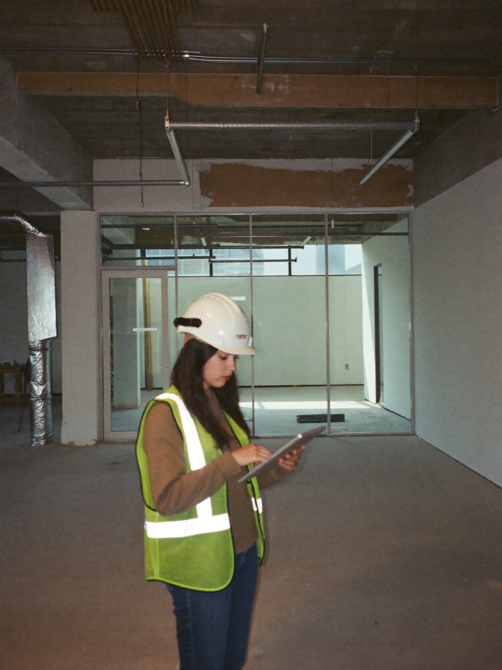 Engineer in a hard hat and safety vest documenting an unfinished commercial interior with a tablet, exposed ceiling and glass curtain wall behind her