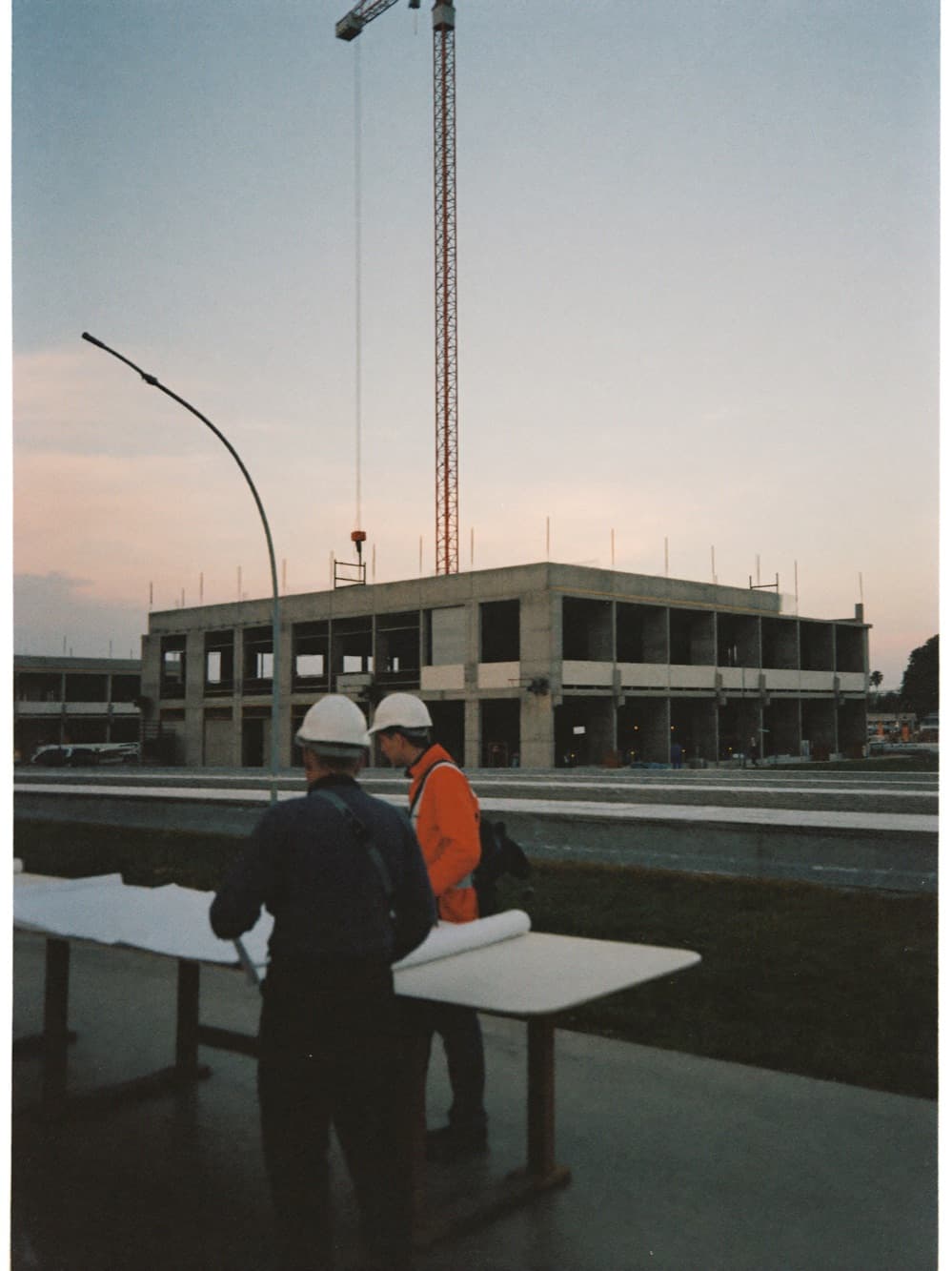 Project manager and superintendent reviewing drawings at a plan table in front of an active mid-rise construction site at dawn