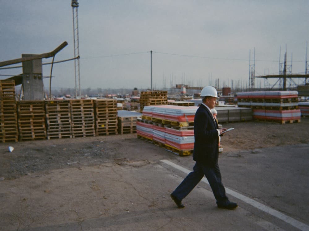 Superintendent in a hard hat walking an active construction site with a tablet, pallets and scaffolding behind him