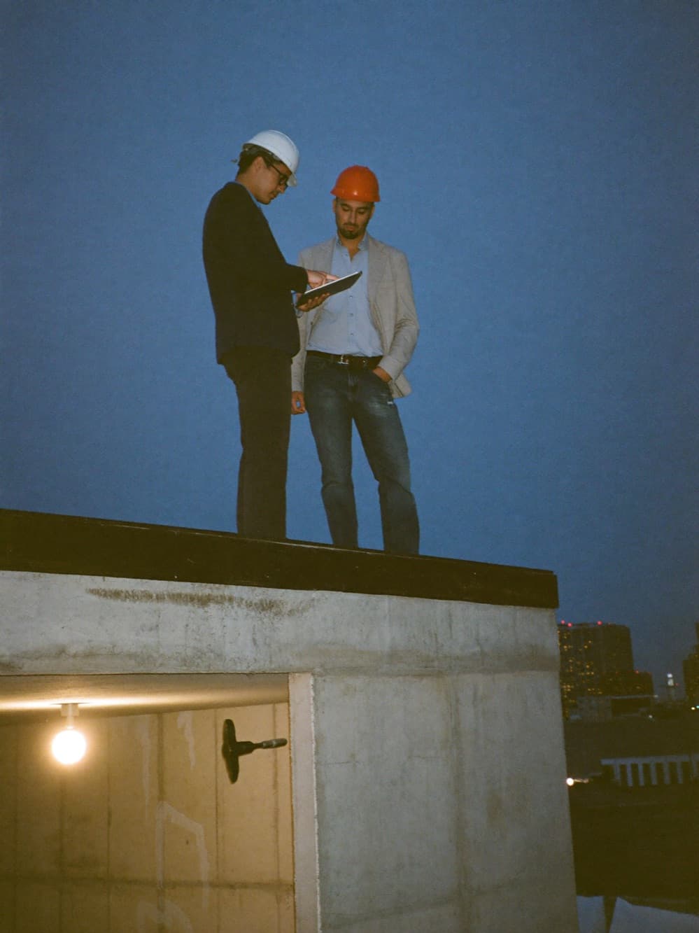 Developer and general contractor reviewing progress on a tablet on an upper floor of a concrete building shell at dusk, city skyline beyond