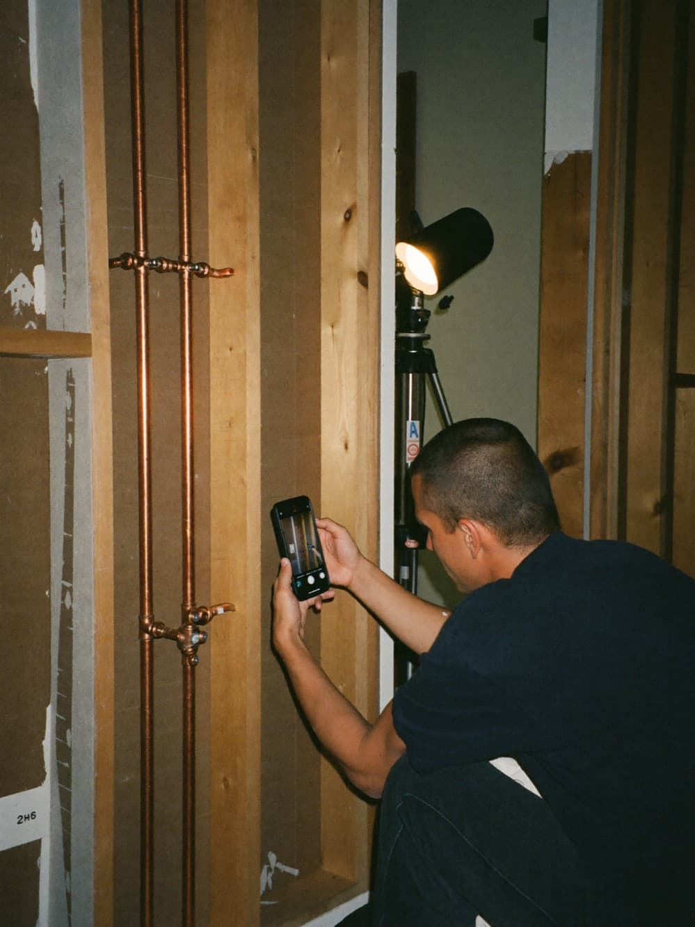 Plumber photographing a completed copper rough-in with his phone in an unfinished wall, work light glowing behind him